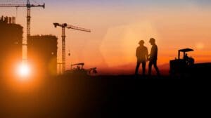 2 construction workers in silhouette with cranes and equipment in the background, a sunset sky.