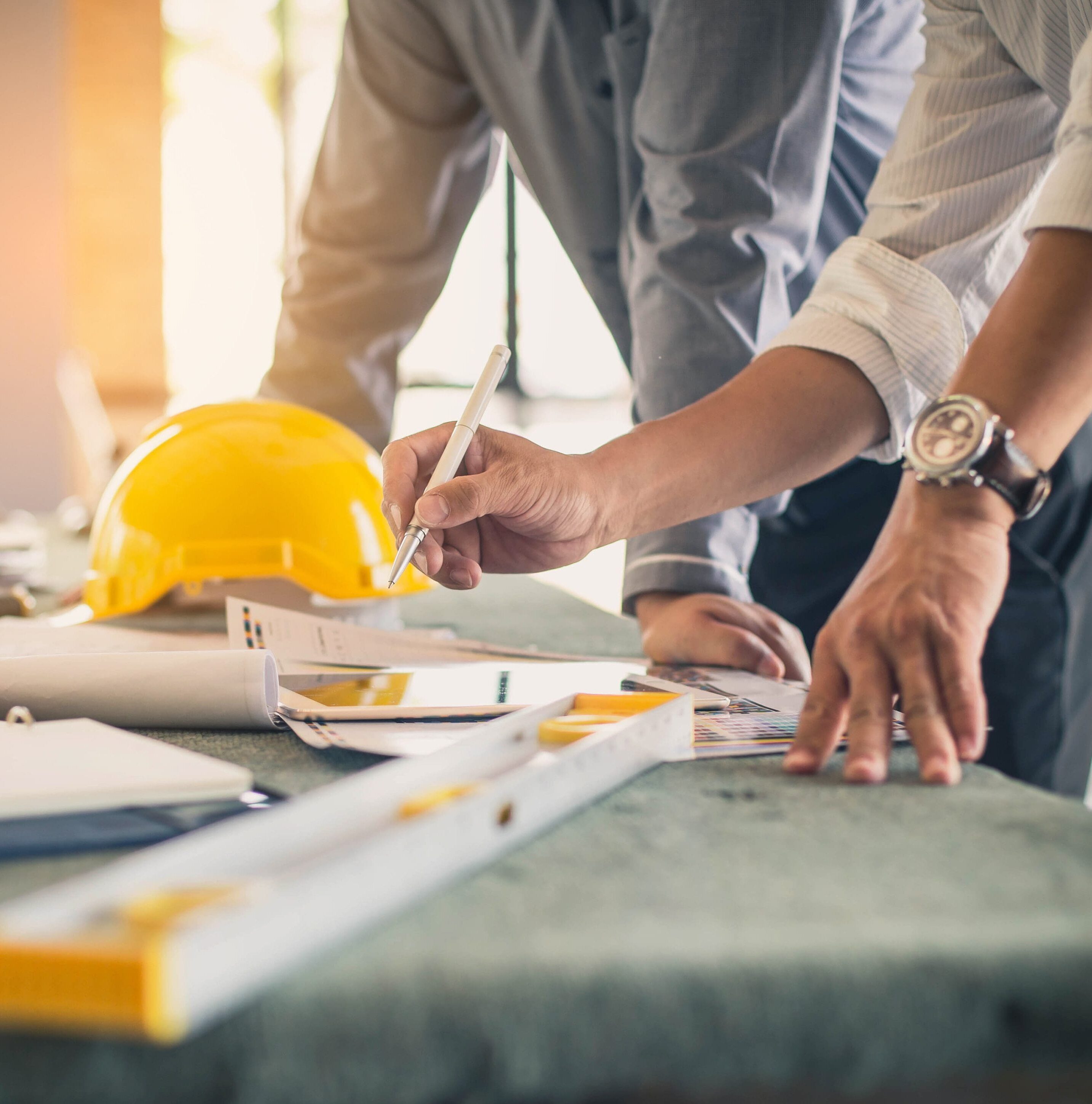 Two site engineers standing at a desk, discussing. A yellow hard hat, papers, and equipment are on the desk.