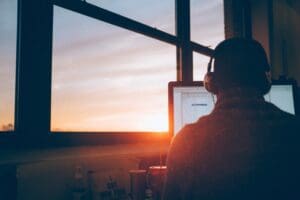 a picture of someone on their laptop facing away from the camera, sunset background through the window, they are in silhouette