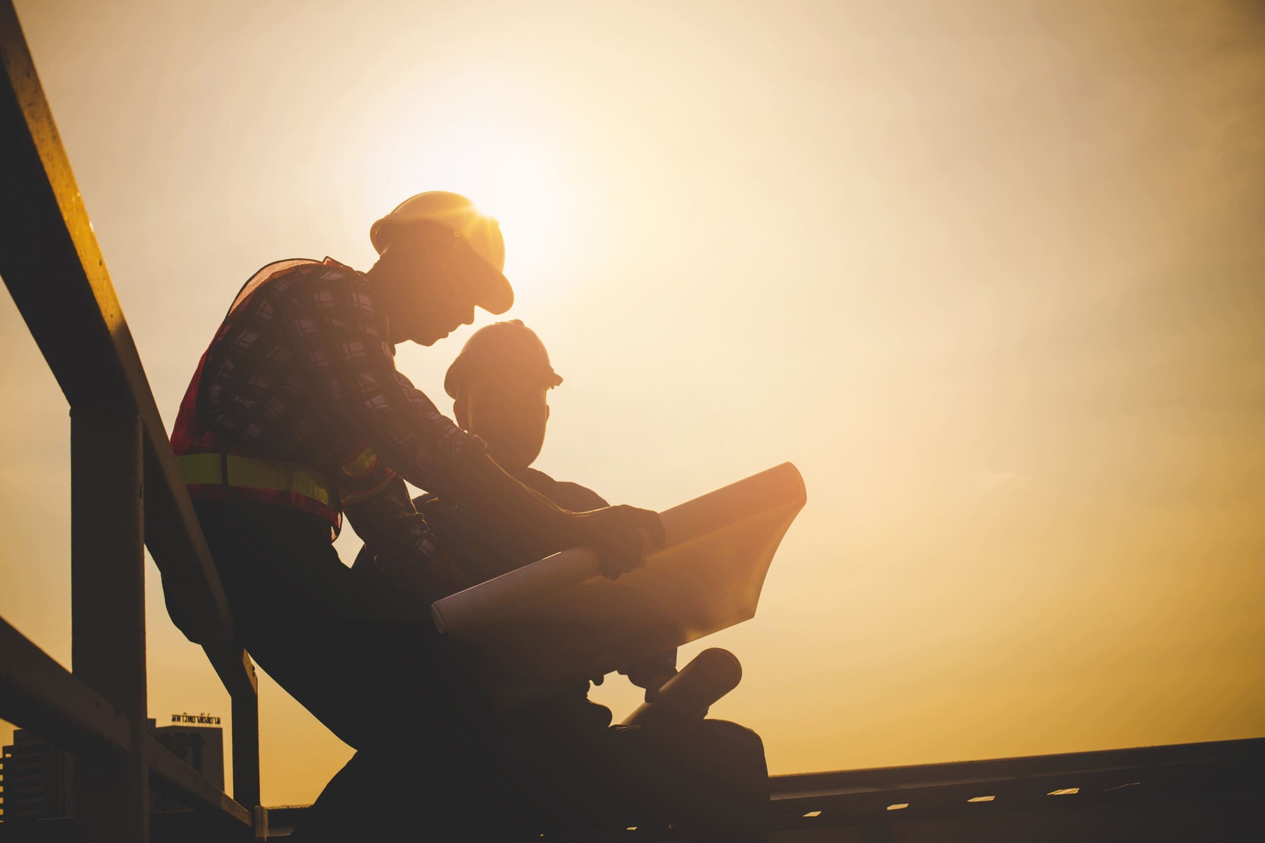 Two construction workers in silhouette examining a blueprint, with a bright yellow sky in the background.