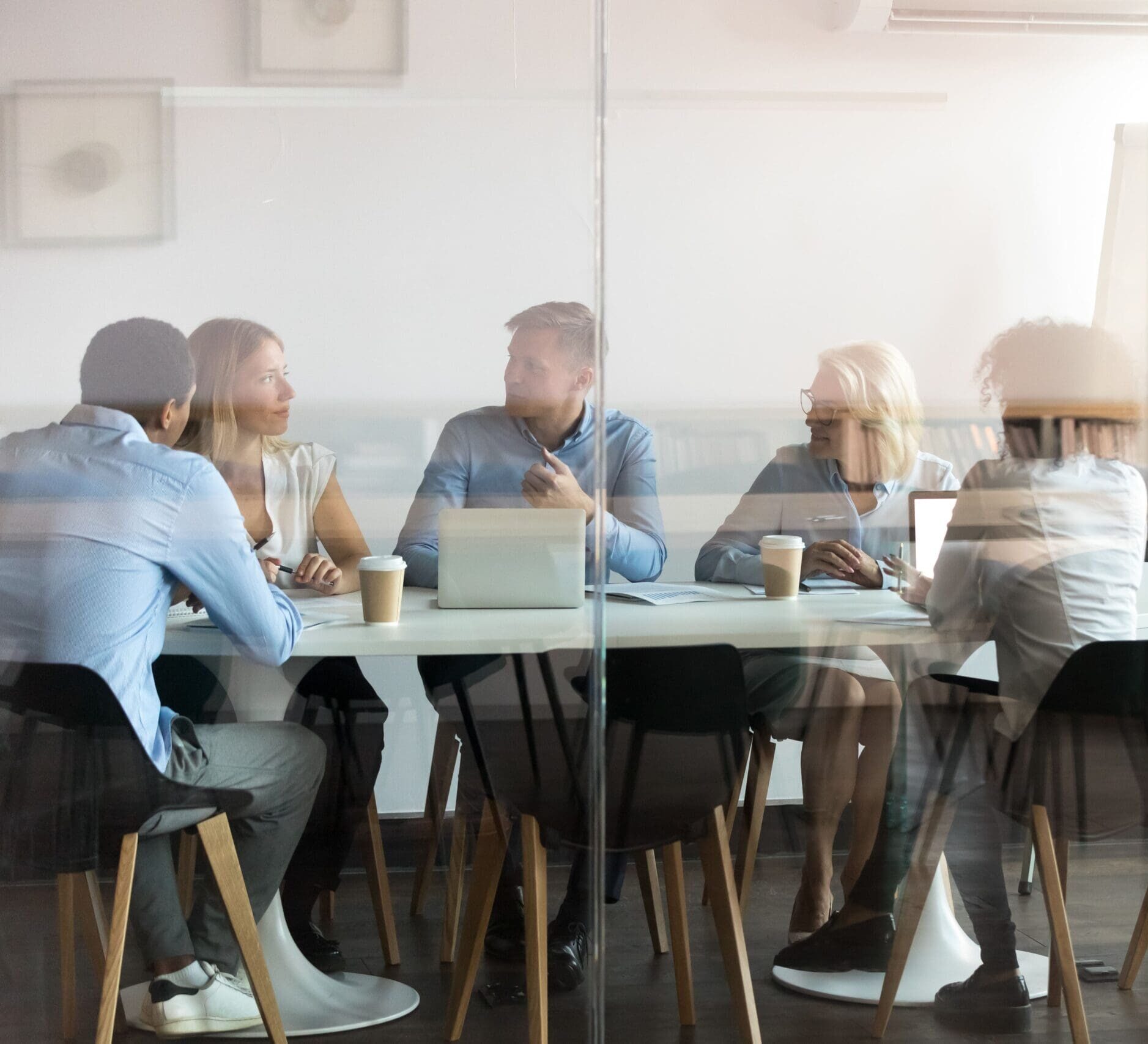 View through glass: business people seated at a table with laptops and coffee, chatting together.
