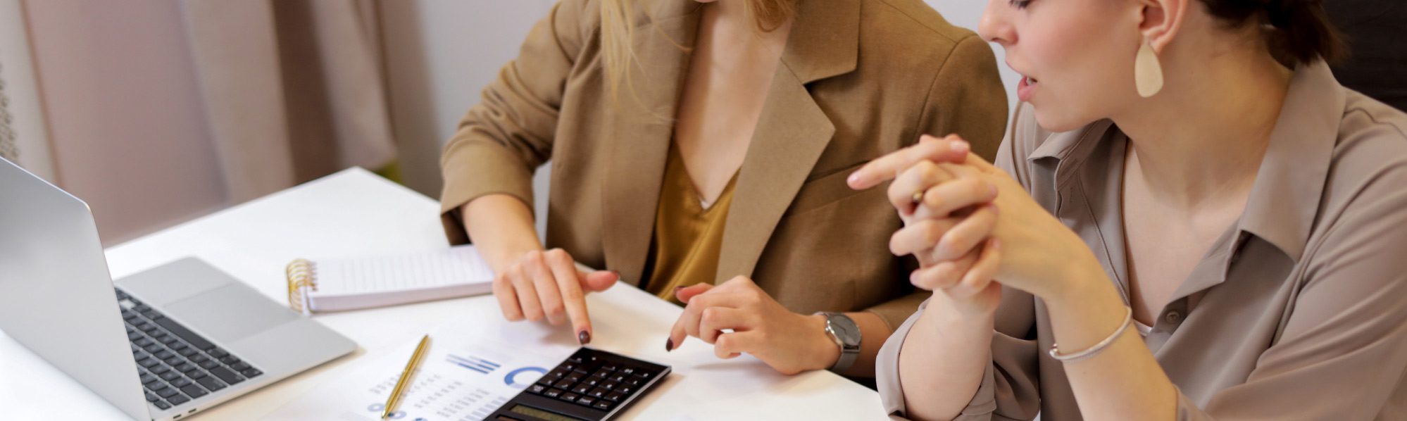Two businesswomen at a desk, one using a calculator, paperwork spread out, in a modern office setting.