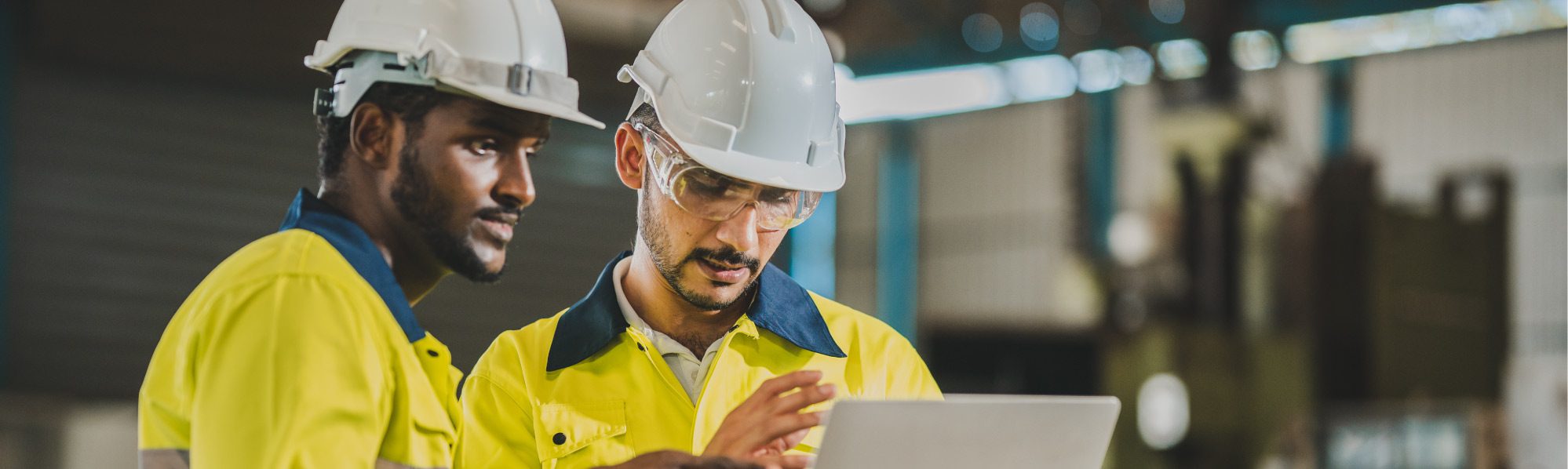 Two contractors in PPE with white hard hats standing, talking, and looking at a laptop.