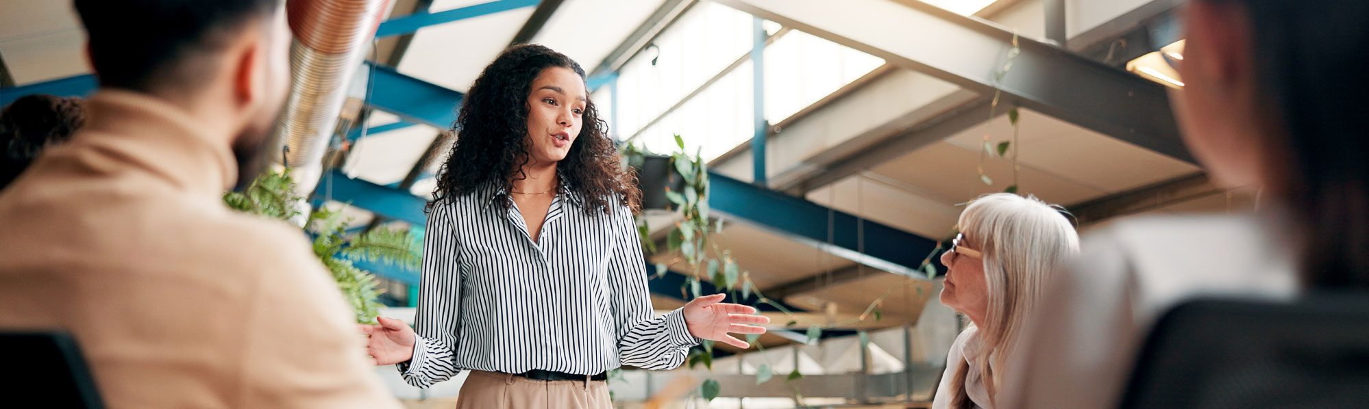 A group of businesspeople in a bright office, some seated, listening to a female speaker.