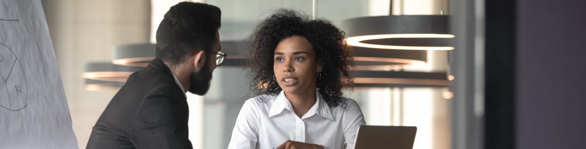 A business man and woman sat at a table with a laptop out, having a conversation.