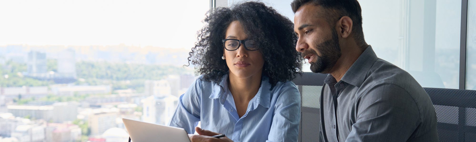 Two colleagues sit at a desk discussing information on a laptop in a bright office overlooking a city.